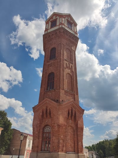 A tall, red-brick water tower stands prominently against a bright blue sky with scattered white clouds in the city of Staraya Russa, Russia. The tower, identified as the Staraya Russa Water Tower, features a broad, multi-faceted base constructed of a lighter, possibly stone or concrete, material. Above the base, the main shaft of the tower is made of red brick, characterized by decorative brickwork including recessed arched panels and horizontal banding.

At the lowest visible level of the brick shaft, there is a large, arched window with a dark frame and intricate multi-pane glazing. Higher up the shaft, several more tall, narrow arched windows with dark frames are present, interspersed with decorative arched recesses without glass. The tower tapers slightly before transitioning to its upper section.

The top segment of the tower is an octagonal observation level, distinguished by a contrasting color scheme of white and orange-red trim. This level features multiple windows and an exterior balcony with railings. A pointed roof, possibly made of metal, caps the entire structure.

To the left of the tower, partial views of lush green tree foliage, a black lamppost, and segments of two light-colored residential buildings with red roofs are visible. To the right, a denser expanse of green tree canopies suggests a park or tree-lined street, with indistinct architectural elements visible through the foliage in the distance. The sky is predominantly clear blue, adorned with scattered white cumulus clouds and some wispy cirrus clouds, indicative of daytime conditions. No people or specific actions are discernible within the frame.