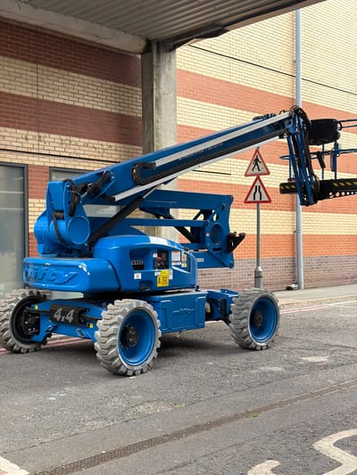 A blue Genie boom lift, identified by the "4x4" marking on its chassis, is parked on an asphalt road in front of a brick building in South Ockendon, United Kingdom. The building features alternating bands of red and cream bricks. Two triangular traffic signs are visible behind the boom lift: one depicting a pedestrian crossing and another indicating a narrowing of the road ahead. The boom lift has large, rugged tires and a visible control panel on its base. A section of a concrete column and an overhead canopy are also present in the scene.