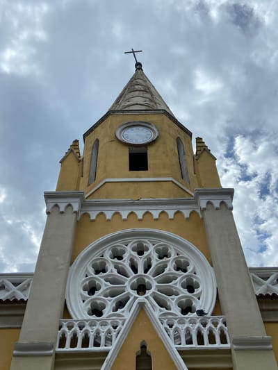 The image shows a low-angle upward view of the tower of the Iglesia Nuestra Señora del Carmen in San José de Barlovento, Venezuela. The tower is painted yellow and has white trim. The spire is topped with a metal cross. The tower has a clock face, a round decorative window, and other architectural details. The sky behind the tower is cloudy.