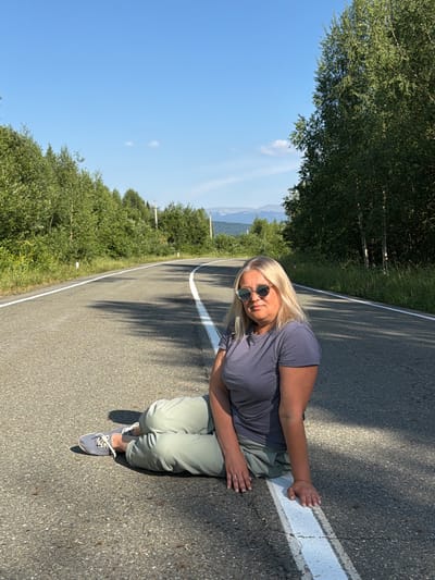A blonde woman with sunglasses is sitting on the side of a paved road. She is wearing a grey t-shirt and light green pants, and blue sneakers. The road has a white line painted on its right side. The woman is looking towards the camera.
The scene is set in a rural area with lush green trees and vegetation lining the road. In the background, mountains are visible under a clear blue sky with a few white clouds. The sun is casting shadows on the road, suggesting it is daytime, likely mid-morning or afternoon. The overall atmosphere appears relaxed and peaceful. There are no visible signs or text in the image.