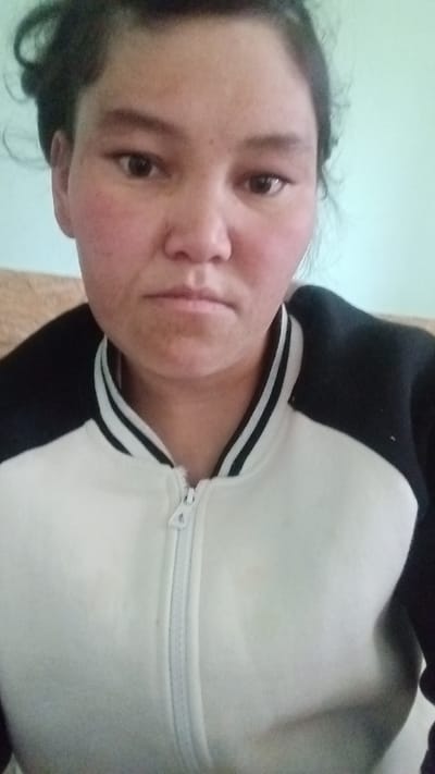 The image shows a close-up, front-facing selfie of a young woman with dark hair and brown eyes. She has rosy cheeks and is wearing a black and white zippered jacket. The background is a solid light blue wall. The photo appears to have been taken indoors, likely in a residential setting. No specific location in Aydarken, Kyrgyzstan is identifiable from the image.