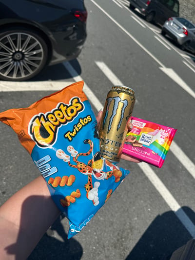 The image shows a person's hand holding a bag of Cheetos Twistos, a Monster Energy Golden Apple drink, and a Ritter Sport Unicorn chocolate bar. The hand is positioned in front of a background of parked cars and a striped asphalt surface. The image appears to be taken outdoors. Some text in the image suggests the location may be Varazze, Italy.