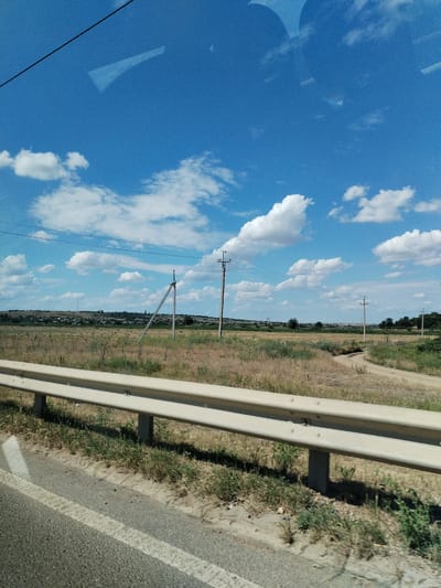 The image depicts a rural roadside scene under a bright blue sky with scattered white clouds. In the foreground, a beige metal guardrail runs along the edge of a paved road, marked with a white line. Beyond the guardrail, a dry, grassy field stretches towards a distant horizon. The field is dotted with numerous hay bales.
Several utility poles with wires are visible across the landscape, suggesting the presence of infrastructure. The landscape is gently rolling, with a small settlement of houses nestled in the distance on a low hill. A dirt road curves through the field.
The scene appears to be set during the daytime, likely late morning or early afternoon, given the bright sunlight and clear sky. The weather seems fair and warm, with the dry vegetation indicating a sunny period. The overall impression is one of a quiet, rural environment in Ukraine. There is no visible text in the image.