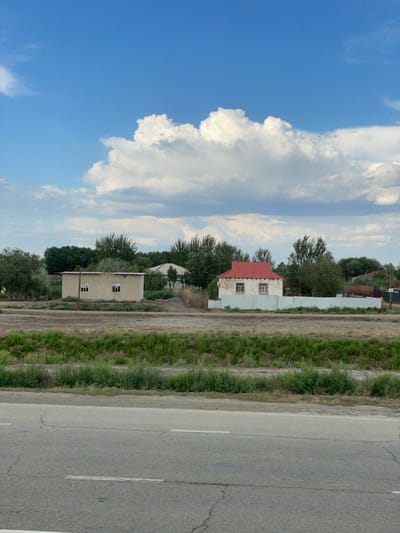 In Qtay, Uzbekistan, a rural landscape unfolds under a partly cloudy blue sky. In the mid-ground, a beige, rectangular building with two windows sits on a dirt road, possibly a dwelling or small structure. To its right, a white house with a red tiled roof and two windows is partially visible, surrounded by a pale metal fence. Beyond these structures, dense green trees form a backdrop, with more scattered trees and vegetation appearing further in the distance. A strip of green grass and brush runs across the foreground, bordering a cracked asphalt road with white lane markers. The scene conveys a sense of quiet, residential life in a semi-arid environment.