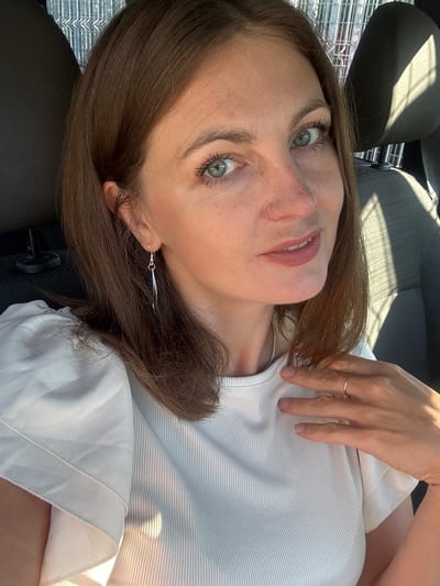 This is a close-up, medium shot selfie of a woman in her late 30s or early 40s. She has medium brown hair, fair skin, and striking light blue eyes. She is wearing a white ribbed top with puffed sleeves and a silver dangle earring. A thin gold ring is visible on her right ring finger. The woman is in the driver's seat of a car, evidenced by the car seat behind her and the metal grating of what appears to be a parking garage or similar structure in the background, suggesting a setting within the city of Lavans-lès-Saint-Claude, France. The lighting suggests it is daytime, with sunlight casting shadows.