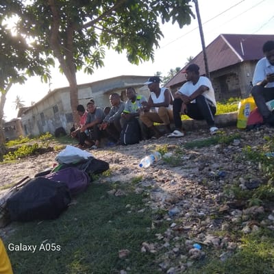 The scene is located at the coordinates -5.726 latitude, 39.301 longitude, which is in Nungwi, Tanzania. Seven young men sit on the ground under a tree, beside a concrete building. The building has a brown, tiled roof. The men are wearing casual clothing. They are talking and interacting with each other. Some are wearing backpacks. Several items are scattered on the ground, including a plastic bottle, a backpack, and a plastic bag. The ground is a mix of dirt, gravel, and grass. The scene is located in a rural area, as there are no cars or other vehicles present. There is a large, green tree providing shade for the men. The sun is shining, casting shadows from the tree and buildings. The background shows other structures in the area, including a small, single-story building.
