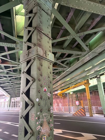An eye-level, medium shot captures the underside of a green painted, multi-level railway overpass in Chiyoda, Japan. The foreground is dominated by a large, riveted steel pillar, also painted green, adorned with various graffiti tags and stickers. The pillar's structure incorporates a diagonal latticework design. Behind the pillar, a complex network of green and purple painted steel beams and girders forms the overpass structure, with a light reflecting off some of the surfaces. In the background, a yellow gantry crane is visible, along with a red brick wall covered in graffiti. The ground level features a paved road with white lane markings, including arrow indicators. A striped concrete barrier, a yellow pole with black stripes, and a white metal fence are also present. The overall scene suggests an urban environment with active infrastructure.