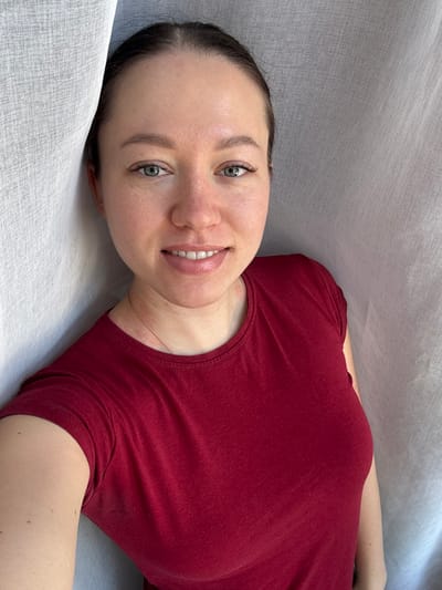 In Gamovo, Russia, an adult female is captured in a close-up, chest-up portrait indoors. The subject has light skin, light-colored eyes, and dark hair pulled back. She is looking directly at the camera with a smile, revealing her upper teeth. She wears a solid, short-sleeved, round-neck red t-shirt, with a thin, light-colored necklace visible around her neck. The background consists of a light-colored, possibly white or off-white, wrinkled fabric, consistent with drapes or curtains, presenting a soft, diffused lighting environment.
