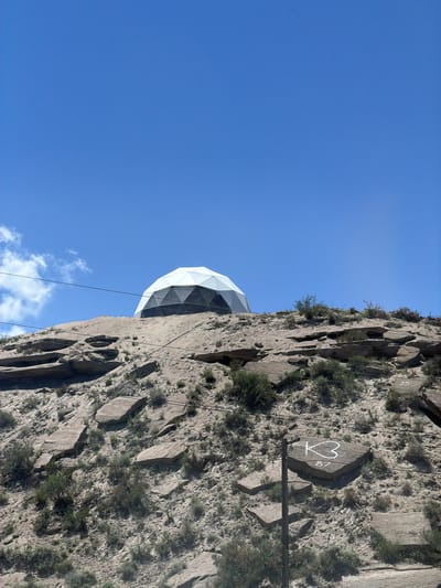 The image shows a geodesic dome structure situated on a rocky, arid hillside under a clear blue sky. The dome is white and grey, with a segmented pattern. The hillside is covered with sparse vegetation and large, flat stones. In the foreground, a rock with the markings "K3" and "87" is visible, alongside a wooden post and power lines. There are no people or animals in the image. The overall impression is of a remote, possibly scientific or observational facility. The specific location in Tong, Kyrgyzstan, is not discernible from the visual information alone.