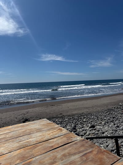 A sunny beach scene is captured with a clear blue sky streaked with wispy clouds. Ocean waves break along a sandy shore, adjacent to a pebble-covered area. In the foreground, a wooden boardwalk provides a sharp contrast to the natural elements, suggesting a constructed pathway alongside the beach.