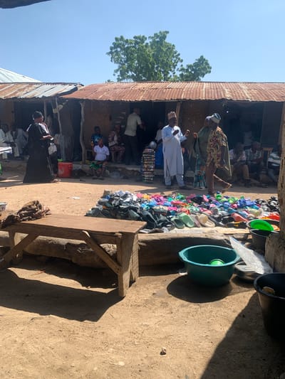 The image depicts an outdoor market scene in Dull A, Nigeria, with a clear blue sky and bright sunlight, suggesting it is daytime. The scene is characterized by rustic, corrugated metal structures and dirt ground. Several people are present, some appearing to be vendors and others shoppers. One man in a light blue garment and cap is interacting with another man wearing a patterned brown and gold robe and a cap. A large collection of colorful sandals and flip-flops are displayed for sale, neatly arranged along a walkway. Green plastic tubs are also visible, one prominently placed near the footwear display and another in the foreground. The overall impression is of a bustling, informal marketplace. The image is oriented vertically, with the sky and a tree visible on the left and the market activity filling the rest of the frame.