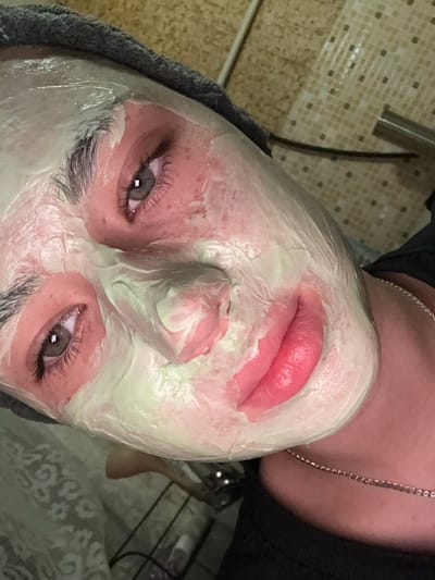 The image is a close-up, low-angle shot of a person's face covered in a pale green face mask, with a dark gray towel wrapped around their head. The person's eyes are open and looking towards the camera. The skin around the eyes and cheeks appears red. The lips are a bright pink color and are slightly parted. A thin silver chain is visible around the person's neck. The background consists of a tiled wall and a patterned cloth. The image is likely taken indoors. There is no information in the image to suggest the location is in the city of Rezh, Russia.