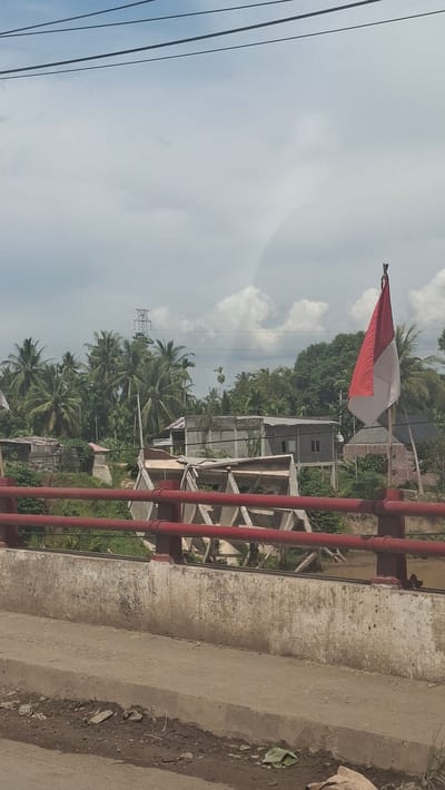 The image is a vertical, eye-level, outdoor shot taken on a cloudy day. In the foreground, a concrete barrier separates the viewer from a red-railed bridge. Beyond the bridge, a river flows, and a broken structure, possibly a water wheel, is partially submerged in it. The background reveals a lush landscape with numerous palm trees and other vegetation. Several buildings, some appearing to be under construction or in disrepair, are visible among the trees. A tall electrical tower stands in the distance. A red and white Indonesian flag is prominently displayed on the right side of the frame, attached to the railing of the bridge. The scene appears to be in a rural or semi-rural area.