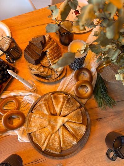 The scene depicts a wooden table laden with various food items. A large round wooden tray holds numerous pancakes, arranged in a circular pattern. Nearby, a smaller wooden tray contains an assortment of sliced bread, including dark and light varieties. Several bagels are positioned near the pancakes and bread. A plate of dark olives is also visible. The table is decorated with a white, textured cloth, adding to the visual appeal. Candles in glass holders and dark-colored glasses are scattered around the table. A vase with greenery is placed on one side, along with a pine sprig. The presence of pancakes, bagels, and olives suggests a possible brunch or celebratory meal. The scene's setting appears to be in Марусино, Russia.