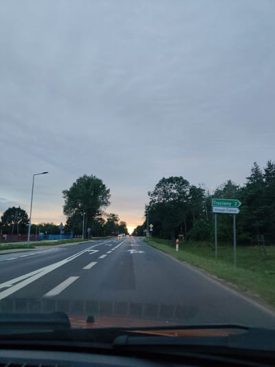 The image captures a view from inside a vehicle driving on a road, likely a highway or a main road, at dusk. The sky is overcast with pale blue and grey clouds. The road has multiple lanes with white markings, indicating traffic flow. On the right side of the road, a green directional sign points to "Trzciany 2" and "Urząd Celny" (Customs Office). Trees line the sides of the road, with a dense forest on the right and more scattered trees and some buildings on the left. The sun is setting on the horizon, visible as a bright, diffused light behind the trees in the distance, casting a warm glow on the sky. Several cars are visible on the road ahead, their headlights on. The foreground shows the dashboard and windshield of the vehicle, partially obscuring the view. The scene appears to be in a rural or semi-rural area, possibly on the outskirts of a town or city. Given the directional sign, the location is likely near Trzciany and a customs office, within the vicinity of Suchocin, Poland.