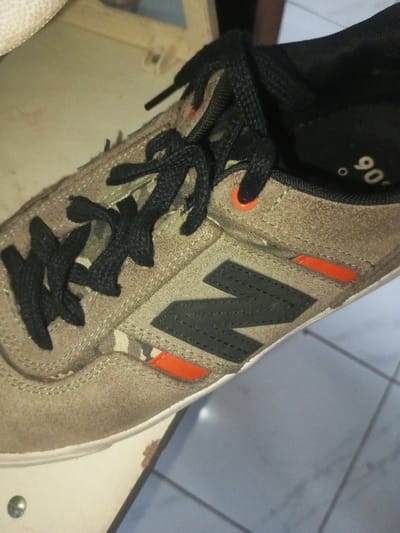 A close-up showcases a tan New Balance sneaker. The shoe features black laces, a black "N" logo on the side, and small orange accents. The material appears to be suede or a similar soft texture. The sole is white. Visible inside the shoe is a black insole with a "90" marking. The sneaker sits on a surface that is partially visible and scuffed.