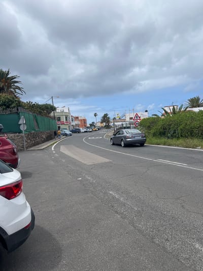 The image depicts a street scene in La Aldea de San Nicolás, Spain. A grey car is driving on a road with a speed limit of 30 km/h indicated by a circular road marking. There are several parked cars to the right side of the road with the tail lights of a red and white car visible in the foreground. Buildings and palm trees line the street. The sky is overcast with some patches of blue visible. A triangular road sign is also visible on the side of the road.