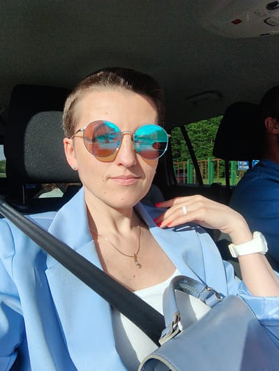 A female individual is seated in the passenger seat of a vehicle, in the Негарэльскі сельскі Савет, Belarus, captured in bright daylight. She has short, dark hair, wears large, round, gold-framed mirrored sunglasses that reflect a bright blue sky, orange tones, and elements resembling a road and vehicles. She is dressed in a light blue blazer over a white ribbed tank top. A thin gold chain with a small pendant is visible around her neck. Her left hand rests on her chest, displaying a light pink silicone ring on her ring finger and bright red nail polish. A white digital wristwatch is on her left wrist, and a small stud earring is in her left ear. A black seatbelt crosses her chest. Partially obscured on her lap is a light blue handbag. To her right, a male individual's head and shoulder are partially visible; he wears a dark blue shirt. Through the vehicle's window, green trees are prominent, behind which a green and yellow fence is discernible. A blue sign with white Cyrillic text, partially reading "УХОД У ЛЕС" (ENTRANCE TO THE FOREST), is visible in the distance. The car's interior features dark upholstery and a dark headliner.