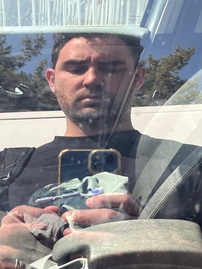A man is sitting in the driver's seat of a car. The man is looking down at a smartphone with a gold case. The phone is reflecting a toothbrush and a towel in the case. He is wearing a black t-shirt. The man is in Kiryat Ye'arim, Israel, at the coordinates (31.806, 35.093). The car is a light gray color. The man's left arm is resting on the armrest. The reflection of trees can be seen in the car window behind the man. The man appears to be in the driver's seat, facing the camera, with a slight, blurred, reflection of the rear view mirror above him. He appears to be wiping the phone screen. The man is the only person in the car. It is a bright day, likely midday. The image is reflected in the car window.