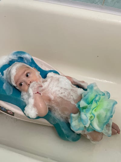 A baby is taking a bath in a bathtub, lying on a blue bath support. The baby is covered in soap suds and has a green and blue bath pouf near their body. The background shows the interior of a bathtub and tiles. The image does not provide any information about the location in the city of Fălești, Moldova.