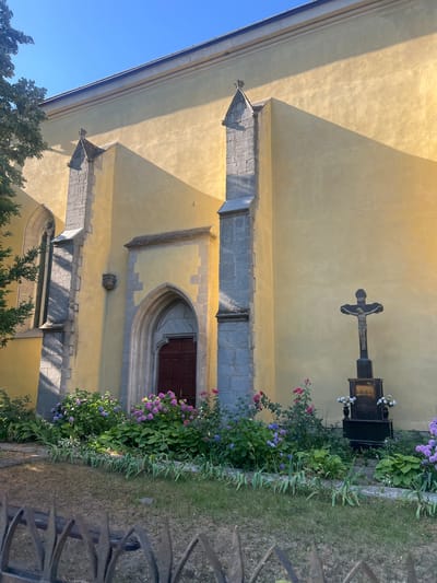 The image shows a side view of a light yellow building, likely a church, with a large arched doorway. The building has gothic architectural elements, including pointed arch windows and stone buttresses. In front of the building, there is a garden with various flowers, including hydrangeas and roses, and a crucifix monument. A metal fence is visible in the foreground. The sky is clear blue. The building is located in the city of Berehove, Ukraine. There are no people visible in the image.