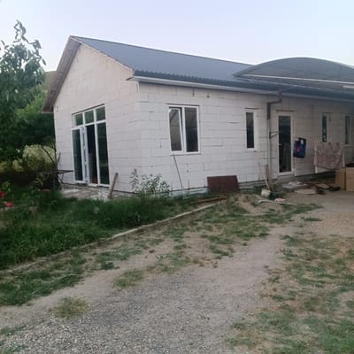 The image shows a house under construction in Prikubanskiy, Russia. The house is made of white cinder blocks and has a dark metal roof. There are several windows and a large glass door. A covered patio extends from one side of the house. The surrounding area is overgrown with grass and weeds, with some gravel and dirt paths. In the foreground, there is a small tree with red flowers and some outdoor furniture. In the background, there are more trees and a hillside. The sky is clear and pale. There are no people visible in the image.