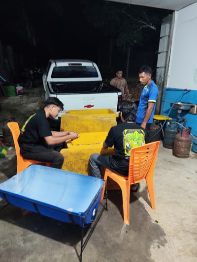 The image depicts a dimly lit outdoor scene, likely in Buket Thagu, Indonesia. In the foreground, a blue plastic tank with a metal frame sits on the ground, reflecting the ambient light. Three individuals are present. Two men, seated on orange plastic chairs, are engaged in an activity, with one appearing to be handling items on a yellow surface behind them. The other man, wearing a blue t-shirt with yellow trim, stands nearby. Behind them, a white pickup truck is parked, with its tailgate down. Several large, yellow rectangular containers are stacked next to the truck. The background is dark with trees and hints of structures, suggesting a semi-rural or industrial area at night. The ground appears to be concrete or a similar paved surface. Two gas cylinders are visible on the right side of the image, one red and one blue.