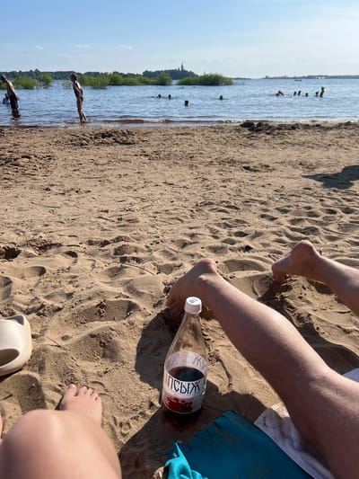 The image depicts a sunny day at a beach, likely on the evening of August 21, 2023, near Ust'ye, Russia. In the foreground, the sandy beach is visible with two pairs of legs lying on it. A bottle of dark red liquid with a label in Cyrillic script is placed on the sand, along with a light-colored sandal and a blue towel. In the mid-ground, several people are in the water, some appearing to swim or wade. The water stretches out to the horizon, where a distant treeline and a building with a spire are visible on the left side. The sky is clear and blue with a few scattered clouds. The overall scene suggests a recreational setting by a body of water.