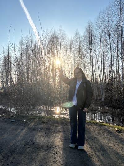 The image captures a young person posing playfully on a dirt path under a bright, sunny sky. The individual, who appears to be female, is positioned slightly off-center, wearing glasses, a dark leather jacket over a light shirt, and dark pants with white sneakers. She is looking towards the camera with a smile, extending her right hand towards the low-lying sun, creating a visual effect where she appears to be holding or interacting with it.

The scene is an outdoor area, likely a trail or forest edge, lined with numerous bare deciduous trees whose branches are silhouetted against the clear blue sky. The sun, low in the sky, is prominently visible on the right side of the frame, casting a warm glow and producing a noticeable lens flare. A distinct white contrail from an aircraft stretches across the upper left portion of the sky.

The ground is a dark, uneven dirt path, with sparse green vegetation and some debris, including a discarded plastic bottle, visible along its edges. The bare trees and the low sun suggest the time of day is either late afternoon or early morning, during late autumn, winter, or early spring. The person's smile indicates a positive and cheerful mood. The location is Novoe Mart'yanovo, Russia. There is no visible text in the image.