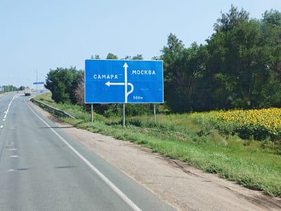 A blue directional road sign in Russian, indicating directions to Samara and Moscow, is positioned on the right side of a two-lane asphalt road. The sign features white text and arrows. To the left, an arrow points straight ahead for Samara. To the right, an arrow curves to the right, indicating Moscow, with "500m" written below it. The road is bordered by a metal guardrail and a grassy embankment. Beyond the embankment, a field of sunflowers is visible, with trees lining the background. The sky is clear and blue. The scene appears to be in a rural or semi-rural area, possibly on the outskirts of a city or town.