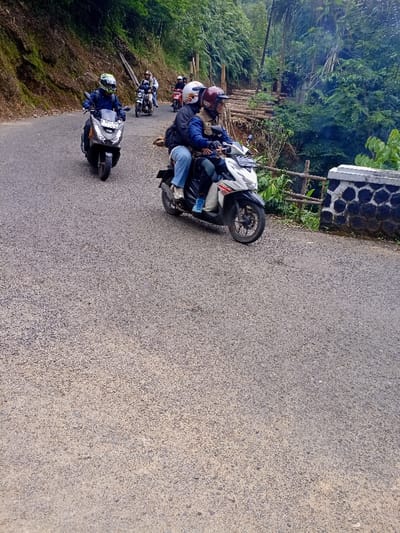 The image shows a paved road with a group of motorcyclists riding on it. There are four motorbikes on the road, with two of them being ridden by two people each. The motorcyclists are all wearing helmets. The road is surrounded by lush greenery, with a stone wall on the right side of the road. There are a few trees and bushes visible. The road leads towards a forested area. Based on the coordinates, this location is most likely a winding road on the edge of the Cisewu District in Garut Regency, West Java, Indonesia.  The scene is a daytime setting, with the sunlight filtering through the trees, making the atmosphere bright. There are no signs of any activity or interaction besides the motorcyclists riding their bikes. The setting is an outdoor rural environment with no evidence of any urban structures.