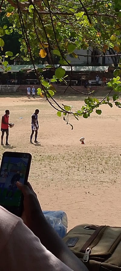 The image depicts an outdoor sports field in Tudor ward, Kenya, viewed from behind foliage. In the immediate foreground, a person's legs are visible along with a dark-skinned hand holding a black smartphone displaying what appears to be a video of people in a similar outdoor setting. A blue plastic-wrapped object and a khaki backpack with a visible zipper are situated next to the legs.
The midground features a predominantly dirt field with sparse green vegetation. Two dark-skinned males are present: one on the left wears a red sleeveless top and dark shorts, holding a white rugby ball; the other, central, wears a blue and white striped jersey with dark shorts and socks, standing and looking downwards. Further to the right, a white rugby ball rests on a reddish-orange kicking tee on the ground. In the background of the field, three additional dark-skinned males in light blue uniforms are standing or walking.
The background beyond the field shows a low concrete structure and several covered shelters with light-colored roofs, possibly spectator stands or auxiliary buildings, with additional structures and trees in the distance. The scene is brightly lit by sunlight, indicating daytime.