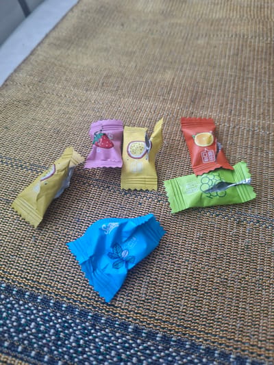 Six individually wrapped candies are scattered on a woven mat. The candies are of various colors, including pink, yellow, red, green, and blue. Each wrapper features a different fruit illustration: a strawberry, passionfruit, orange slice, grapes, and mint leaves. The mat has a textured surface with a pattern of brown, gold, and dark blue threads. The background appears to be a light-colored surface, possibly a couch. There are no people or animals visible in the image.
