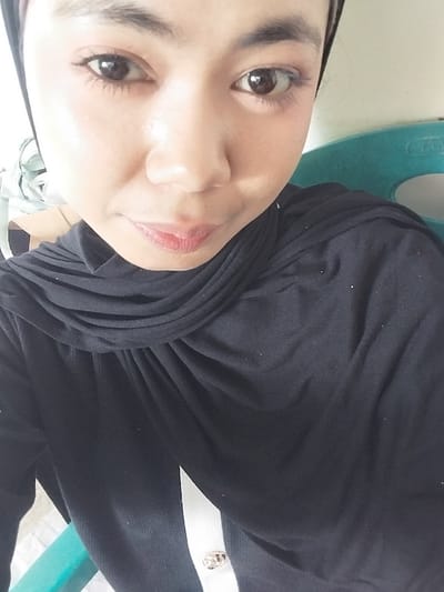 A selfie of a woman wearing a black hijab and a black top is shown. The woman's face is close to the camera, and her eyes are looking directly at the lens. Her lips are slightly parted, revealing a subtle smile. She is wearing light-colored makeup, including eyeliner and mascara. The background is blurred, but a turquoise chair is visible to the right of the woman. The image appears to have been taken indoors, possibly in a home or office setting. The city of Jeppee, Indonesia, is not depicted or implied in this image.