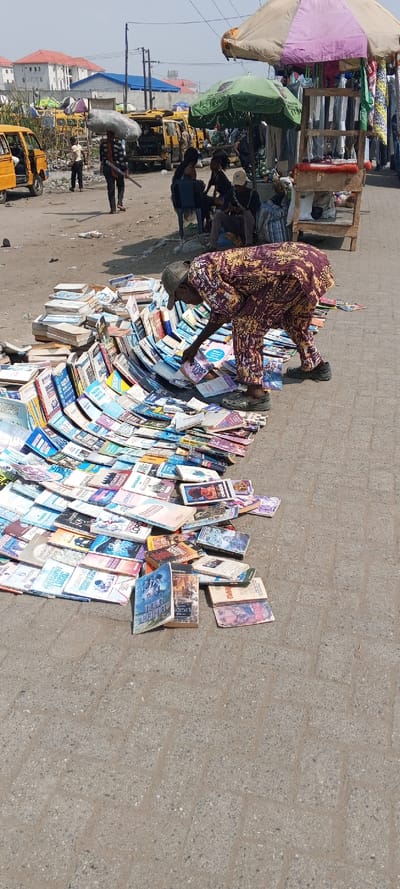 The image displays an active outdoor street market scene in Awjaw, Nigeria, under a clear daylight sky. In the foreground, a large collection of paperback and hardcover books are laid out across a grey interlocking paver block surface, forming multiple scattered piles and linear arrangements. An adult male, dressed in patterned purple, yellow, and red traditional attire consisting of a long-sleeved tunic and trousers, a dark baseball cap, and dark slip-on shoes, is bent over the books, appearing to sort or arrange them with his hands.
To the right of the central figure, a market stall with a wooden shelving unit stands beneath a beige and purple striped umbrella, with textiles hanging behind it. Adjacent, a green umbrella shades several individuals, one of whom, wearing a dark cap, is seated and engaged with a mobile device. In the midground, a yellow commercial minibus with an open door is parked on the left. Further back, another individual walks carrying a large light-colored sack on their head while holding a stick. Multiple other yellow minibuses and various market activities are visible in the background, set against residential or commercial buildings with red and white roofs, utility poles, and overhead wires.