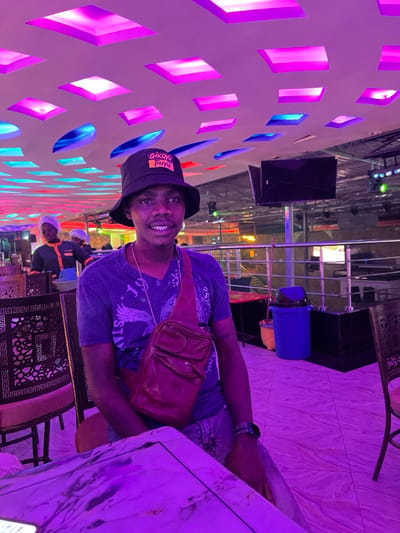 The image captures a young man sitting at a table in what appears to be an indoor entertainment venue or restaurant in Kimorori, Kenya, likely during the evening or night. The scene is illuminated by vibrant artificial lighting, with strong purple, pink, and blue hues emanating from a decorative wall featuring geometric cut-outs.
The main subject is a young man with dark skin, positioned on the right side of the frame. He is wearing a black bucket hat, a blue t-shirt with a white pattern (possibly "MUSTANG"), and light-colored shorts. A brown leather shoulder bag or fanny pack is slung across his chest, and he has a watch on his left wrist. He is looking directly at the camera with a bright smile. The text "GIRL YA HAYU" is visible on the front of his bucket hat.
Behind the man, the background reveals more of the establishment. There are other tables with ornate dark metal bases and light-colored tops, along with chairs. A large black speaker is mounted on a wall, and a blue bin or container is visible further back. Faintly in the deeper background, another person or two in light-colored attire can be seen, possibly staff members. The floor appears to be tiled with a subtle pattern, reflecting the colorful lights. The overall ambiance is modern and lively, typical of a social gathering spot. The photograph is taken at a tilted angle.