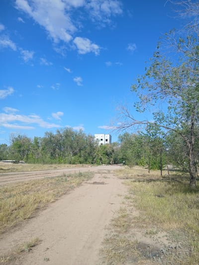 The media file depicts an outdoor scene with a bright blue sky and scattered white clouds. A dirt road runs diagonally through the frame, bordered by dry, yellowed grass and green trees. In the distance, partially obscured by foliage, a white, multi-story building with visible windows can be seen. The image appears to be taken from a slightly elevated perspective, looking down and across the landscape. The overall impression is one of a sunny day in a rural or semi-rural setting, possibly in Kazakhstan given the provided location context. There is no discernible activity or event taking place; the scene is static. No text is visible within the image. The lighting suggests it is daytime, with the sun casting shadows.