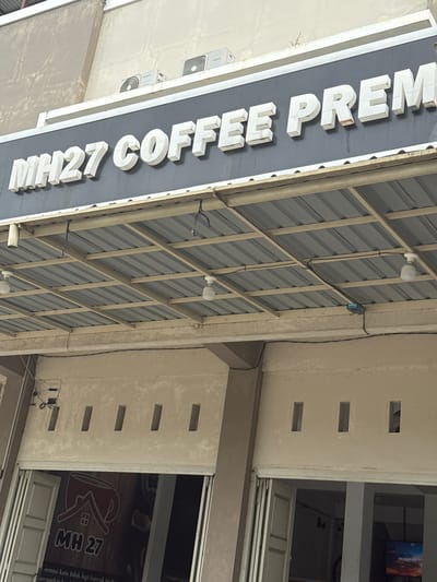The image shows the exterior of a building with a sign that reads "MH27 COFFEE PREM". The sign is made of white 3D letters mounted on a dark background. Above the sign, there are two air conditioning units attached to the wall. A metal awning with a corrugated roof extends over the entrance of the building. The building has a light-colored facade with several rectangular openings. Below the awning, there are open storefronts. In one of the storefronts, a sign with a coffee cup logo and the text "MH 27" is visible. The image appears to be taken during the daytime. The context provided suggests this location is in Meunasahmanyet, Indonesia.