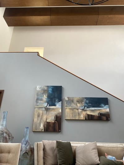 The image depicts an interior view in Santa Cruz Tehuixpango, Mexico. Two abstract paintings hang on a grey wall adjacent to a staircase with a wooden handrail. To the right of the wall, a section of a couch with beige pillows and a person's leg with a bag of ice on the foot is visible. Below, there are two decorative objects resembling blue-painted bottles. To the left, a wooden structure, possibly a shelf or closet, stands against a white wall. A rectangular opening, possibly a small window or niche, is visible higher up on the wall.