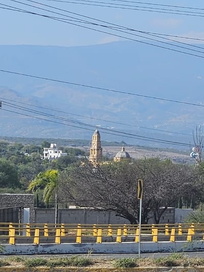 A cityscape features a tall, yellow church with a distinctive dome and spire in the distance, against a backdrop of blue mountains and sky. Utility lines crisscross the foreground. A roadside barrier painted in yellow stands alongside a paved road. Trees and other buildings are scattered throughout the scene.