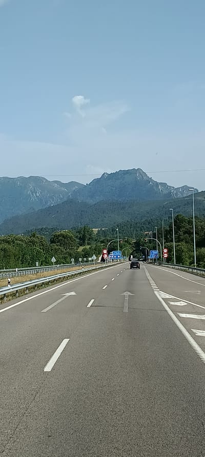 This is a photograph taken from a moving vehicle on a highway, looking ahead and slightly to the left. The highway has multiple lanes, marked with white lines and directional arrows. The road surface is asphalt. On the left side of the highway, there is a steep, rocky mountain covered in lush green trees. The sky is a clear, pale blue with a few wispy white clouds.

Along the side of the road, there are traffic lights and road signs. One set of traffic lights shows a red signal. There are also blue rectangular signs with white symbols and red circular signs with a diagonal line. One of these blue signs appears to indicate a toll road or restriction. The overall impression is of a scenic drive on a clear day, possibly in the late morning or early afternoon, given the bright light. The location is identified as Ribadesella/Ribeseya, Spain, which suggests a mountainous coastal region. No people are clearly visible, but there are a few vehicles on the road ahead. The scene conveys a sense of movement and travel.