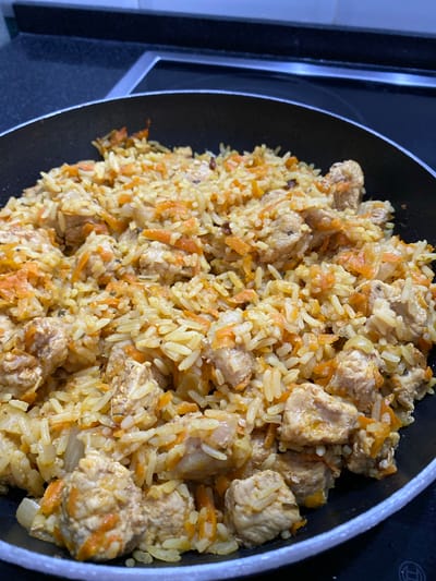A close-up, overhead shot shows a black frying pan filled with a pilaf dish. The pilaf contains rice, diced chicken, and shredded carrots, with visible spices like cumin or coriander. The dish appears to be freshly cooked, with steam gently rising. The frying pan is placed on a black stovetop, suggesting the pilaf is either still cooking or has just been prepared. The background shows the edge of a black countertop and the dark surface of the stovetop, indicating an indoor kitchen environment, likely in Kokkorevo, Russia.