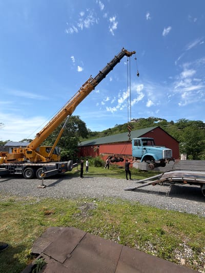 The image depicts an outdoor scene with a large yellow crane lifting the cab of a light blue truck. The crane is positioned on a gravel driveway or road next to a grassy area. Several people are standing near the truck cab, seemingly involved in the lifting operation. A red building with a green roof is visible in the background. The sky is blue with scattered clouds. No indication of the city or area of Kapreshumi, Georgia is visible in the image.
