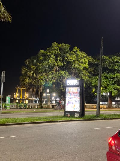 A night-time street view shows a road with a red car's rear visible in the top right. Beside the road, a green strip borders a row of illuminated streetlights. A large sign labeled "painel" is prominent, advertising "carnaval," "centro," and "folia" from "15 a 17". Lush green trees fill the center, contrasting with the dark sky. Below the sign, green bike-sharing stations are visible, adding to the street scene.