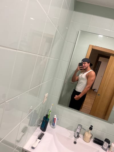 A male individual is depicted in a mirror selfie taken in a bathroom located in Pedrouços, Portugal. The subject, a male with dark hair, wears a dark baseball cap reversed, a white sleeveless top, and dark trousers. He has a gold-colored chain around his neck and is holding a black smartphone in his left hand to capture the image, with his right arm slightly flexed. The bathroom features light blue-green square tiled walls with white grout lines. A white ceramic sink is visible, equipped with a chrome single-lever faucet. On the countertop to the left of the sink are a white-handled hairbrush, a dark blue bottle, a green electric toothbrush, and a clear glass containing a tube of Colgate toothpaste. To the right of the sink, a pump-dispenser bottle with light-colored liquid, a black brush, and other small toiletries are arranged. A white electrical outlet is mounted on the tiled wall to the left. The mirror's reflection reveals a light-to-medium wood-toned door in the background, leading to an interior space with a light-colored floor, where a discarded white garment is visible on the floor.