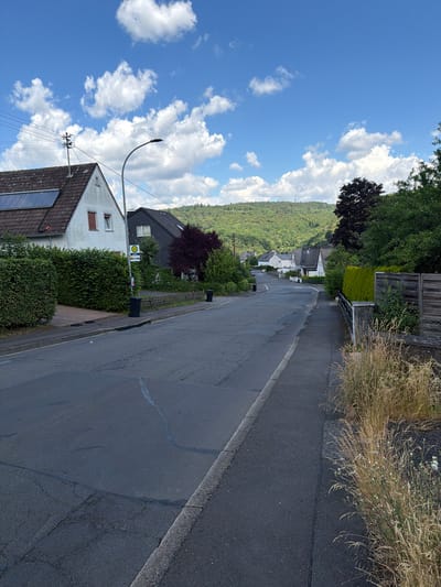 The image depicts a street in Idar-Oberstein, Germany, on a clear day with a blue sky filled with fluffy white clouds. The street is paved and appears to be in a residential area, with houses lining both sides. On the left, a white house with solar panels on its roof is visible, adjacent to a neatly trimmed green hedge. Further down the street, a bus stop sign with a green 'H' symbol indicates public transportation availability. A dark gray house with a sloped roof stands next to the white house. The right side of the street features a sidewalk and a wooden fence separating the properties from the road. Tall, dry grasses and some flowering plants grow along the edge of the sidewalk. In the background, a lush green hillside covered in trees rises up, with a few more houses scattered along its lower slopes. The overall impression is of a quiet, suburban street scene.