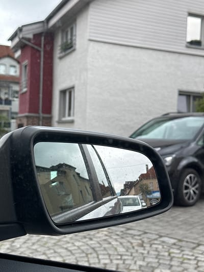 This is a close-up, vertical, slightly angled shot focusing on the side-view mirror of a car. The mirror reflects an urban scene with buildings and cobblestone pavement.

The main subject is the car's side-view mirror, its black casing prominent in the foreground. The mirror itself is speckled with raindrops, suggesting it is raining or has recently rained. Inside the mirror's reflection, we see a building with a red facade and windows, and further back, another lighter-colored building. The reflection also shows parts of other vehicles, including a tire and wheel of a dark-colored car, and a white van or truck. A sliver of the car's interior is visible along the right edge of the frame, including a gray fabric panel.

In the background, out of focus, are the side of a building with vertical white siding and multiple windows, and another building with a red horizontal band. The street surface is visible in the upper right corner, paved with light-colored cobblestones.

The weather appears to be overcast and rainy, indicated by the raindrops on the mirror and the muted lighting. The scene suggests a residential or mixed-use urban area, possibly in Rudolstadt, Germany, as suggested by the location context. No people are visible, and no text is discernible. The overall impression is that of a mundane, everyday scene captured during a rainy day.