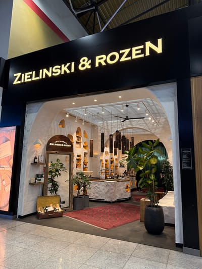 The image shows the exterior of a perfume shop called ZIELINSKI & ROZEN. The shop has a distinctive arched entrance with a rustic, white, textured facade. Inside, there are shelves stocked with various products, decorative lighting fixtures hanging from the ceiling, and plants. A large mirror is visible near the entrance, framed by wood. A red patterned rug is placed in front of the shop, and a suitcase filled with bottles is displayed on the floor to the left. The shop's name is illuminated in gold lettering on a black sign above the entrance. The surrounding environment appears to be a commercial area, possibly an airport or shopping mall, with tiled flooring and other store fronts visible in the background. No people are clearly identifiable in the foreground, though figures can be seen inside the shop in the background. The location is identified as Perepechino, Russia.