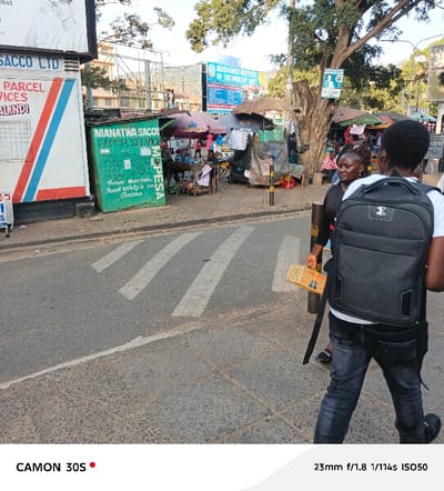 An eye-level, daytime shot shows a bustling street scene in what appears to be Kenya. In the foreground, two individuals walk across a zebra crossing, one with a backpack. Across the street, market stalls are shaded by colorful umbrellas. A vibrant green building advertises "NIANATWA SACCO" with a handwritten message promoting travel safety. Further back, a blue sign for "MACHAKOS INSTITUTE OF TECHNOLOGY" is visible. The backdrop features a hilly landscape and clear sky.