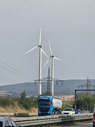 The image captures a view of the A3 Autobahn near Bad Camberg-Kernstadt, Germany. In the foreground, a blue Mercedes-Benz truck with a tanker trailer, identified by the "CEMENTBOUW" logo, is traveling on the highway. To the left of the truck, a silver car is partially visible. Behind the vehicles, a landscape features rolling hills covered in vegetation, with two prominent wind turbines towering in the background. Electrical pylons and overhead power lines are also visible, indicating the presence of significant infrastructure in the area. The sky is overcast with a light gray hue.